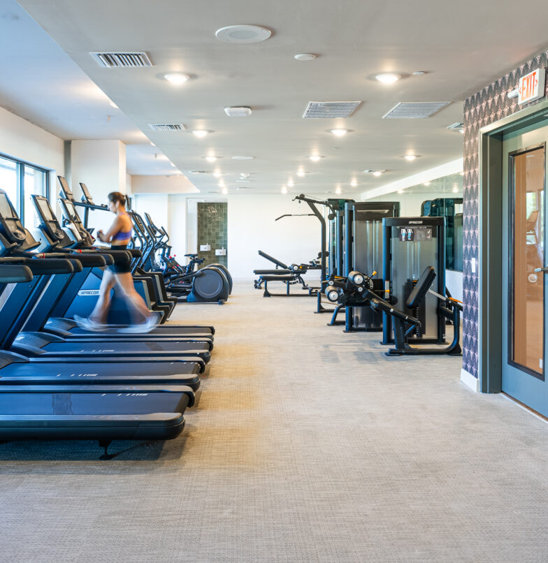 A spacious, bright gym with treadmills, weight machines, and exercise equipment. A person is running on a treadmill near large windows that let in natural light. The gym is clean and modern.