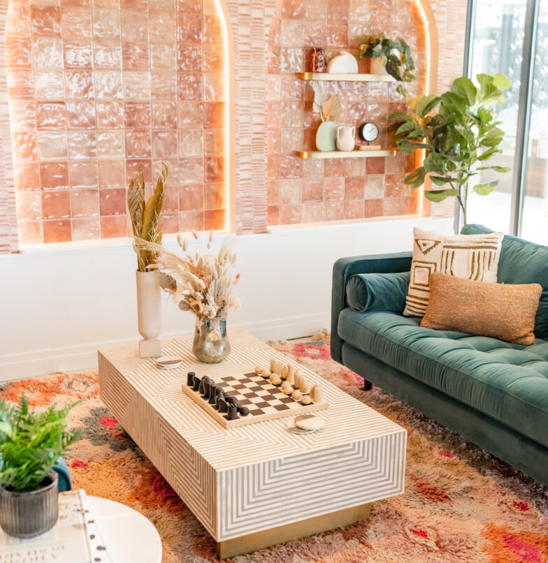 A stylish living room with a green velvet sofa, a textured coffee table with a chess set, dried floral arrangements, a patterned rug, potted plants, and pink tile accent walls with shelves holding decor items.