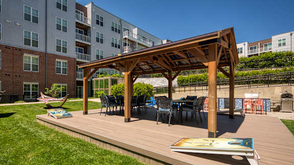 A wooden pergola with chairs and a table is in the foreground of a grassy area with apartment buildings in the background.