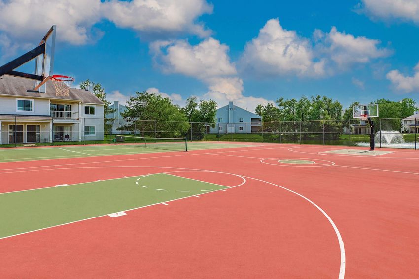 A basketball court with a vibrant red surface, featuring two basketball hoops on opposite ends. The background includes residential buildings and greenery, under a clear blue sky with scattered clouds.