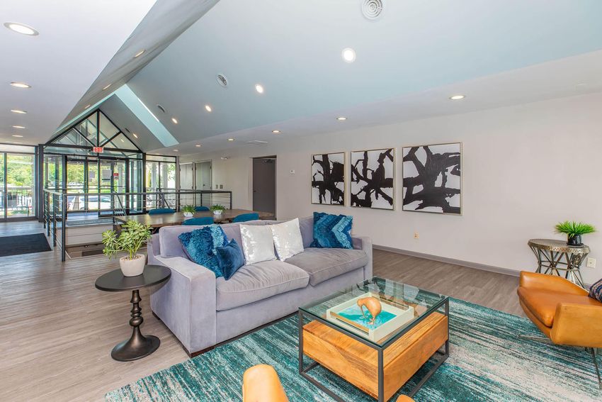 A modern lounge area featuring a gray sofa with decorative blue pillows, a glass coffee table with a decorative bowl, and a wooden side table. The walls are adorned with abstract art, and there is a large glass entrance visible in the background. The flooring is hardwood, complemented by a colorful area rug.
