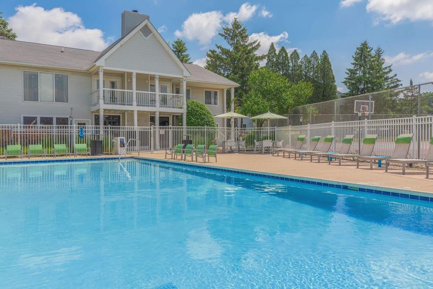 A clear blue swimming pool surrounded by lounge chairs and umbrellas, with a two-story building in the background. Tall trees and a fenced area are visible, under a bright sky with fluffy clouds.