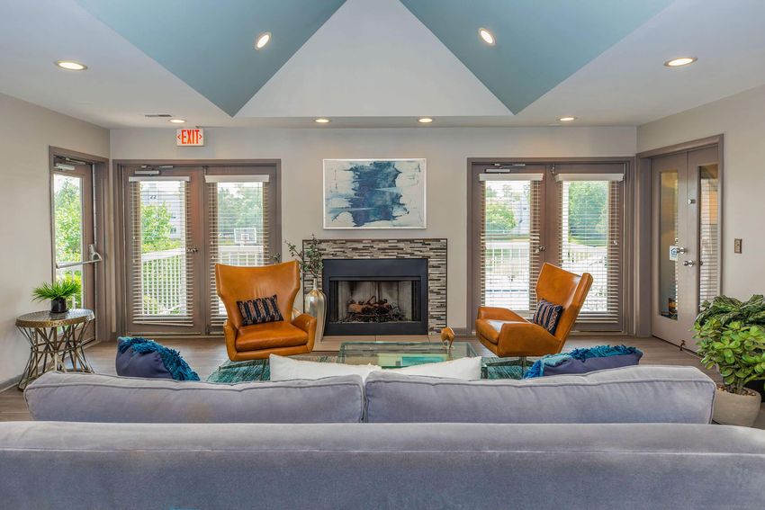 A modern living room featuring a cozy seating area with two orange armchairs facing a fireplace, a gray sofa in the foreground, and large windows with blinds allowing natural light. There's a decorative table, indoor plants, and abstract artwork on the wall, creating a warm and inviting atmosphere.
