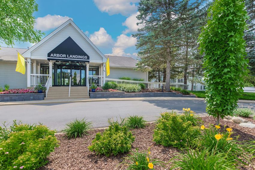 Exterior view of Arbor Landings, featuring a welcoming entrance with glass doors, surrounded by landscaped gardens, colorful flowers, and tall trees under a bright blue sky. Two yellow flags are displayed at the entrance.