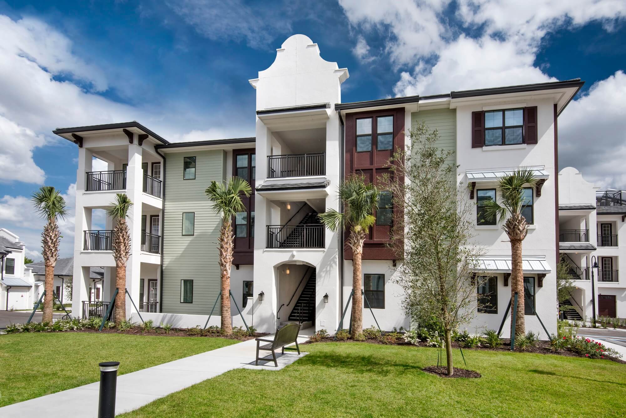 A modern three-story apartment building with white and light green exterior walls, balconies, palm trees, and landscaped lawn under a partly cloudy sky. A bench sits on a walkway in front.