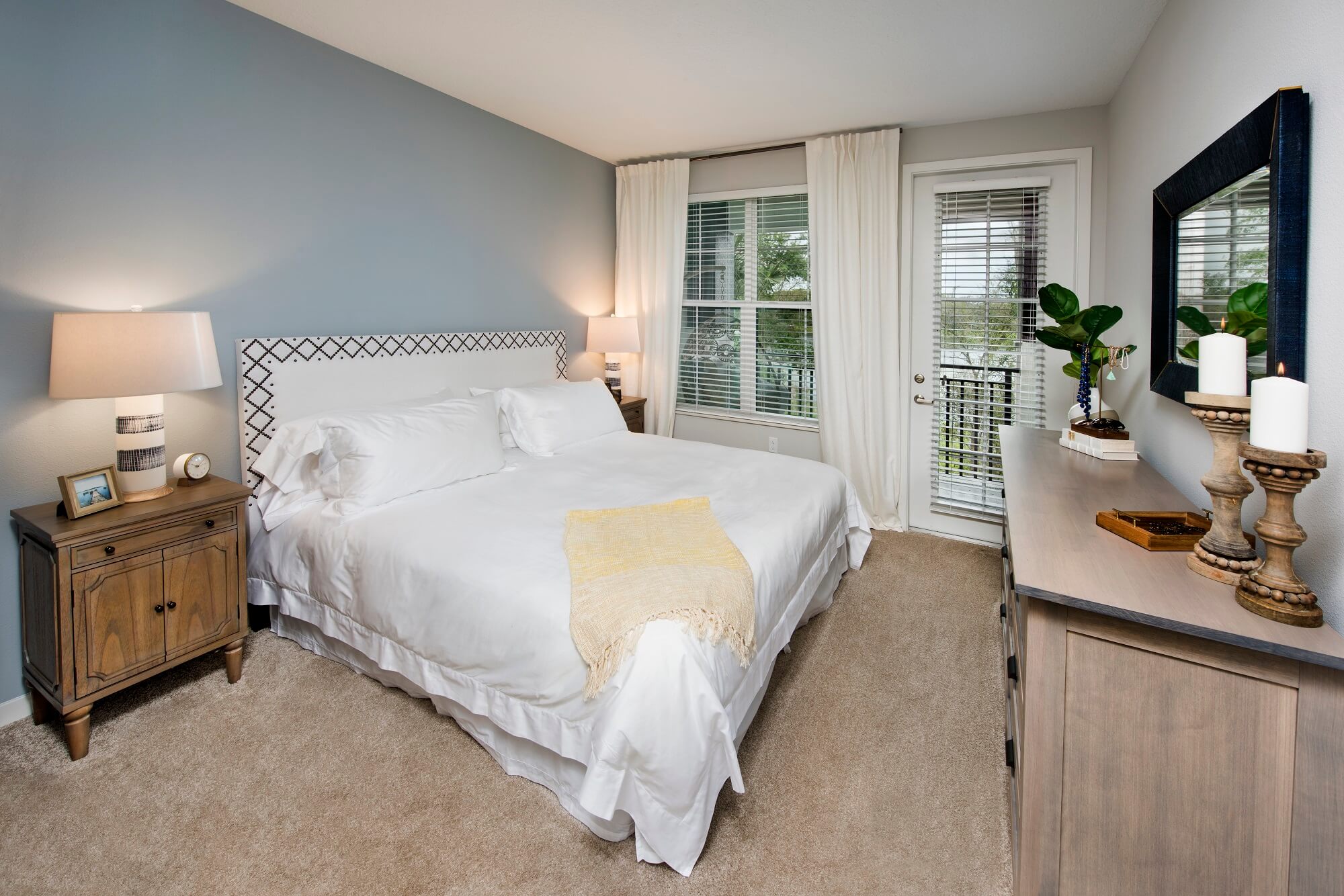 A neatly arranged bedroom with a large bed, white bedding, a yellow throw, two bedside tables with lamps, a dresser with decor, and large windows with white curtains letting in natural light.