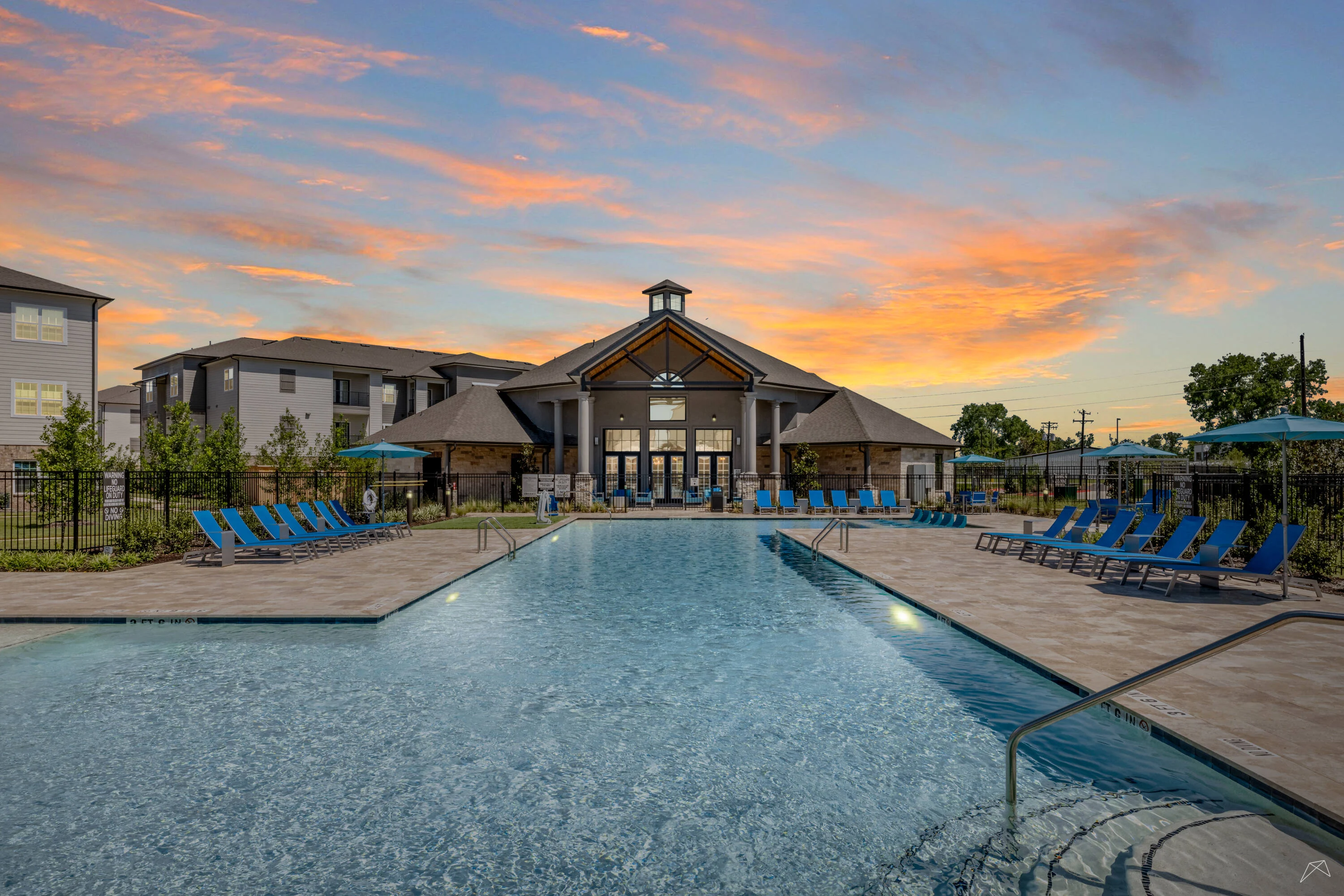 A serene apartment complex with a sparkling pool and blue lounge chairs, set against a vibrant sunset.
