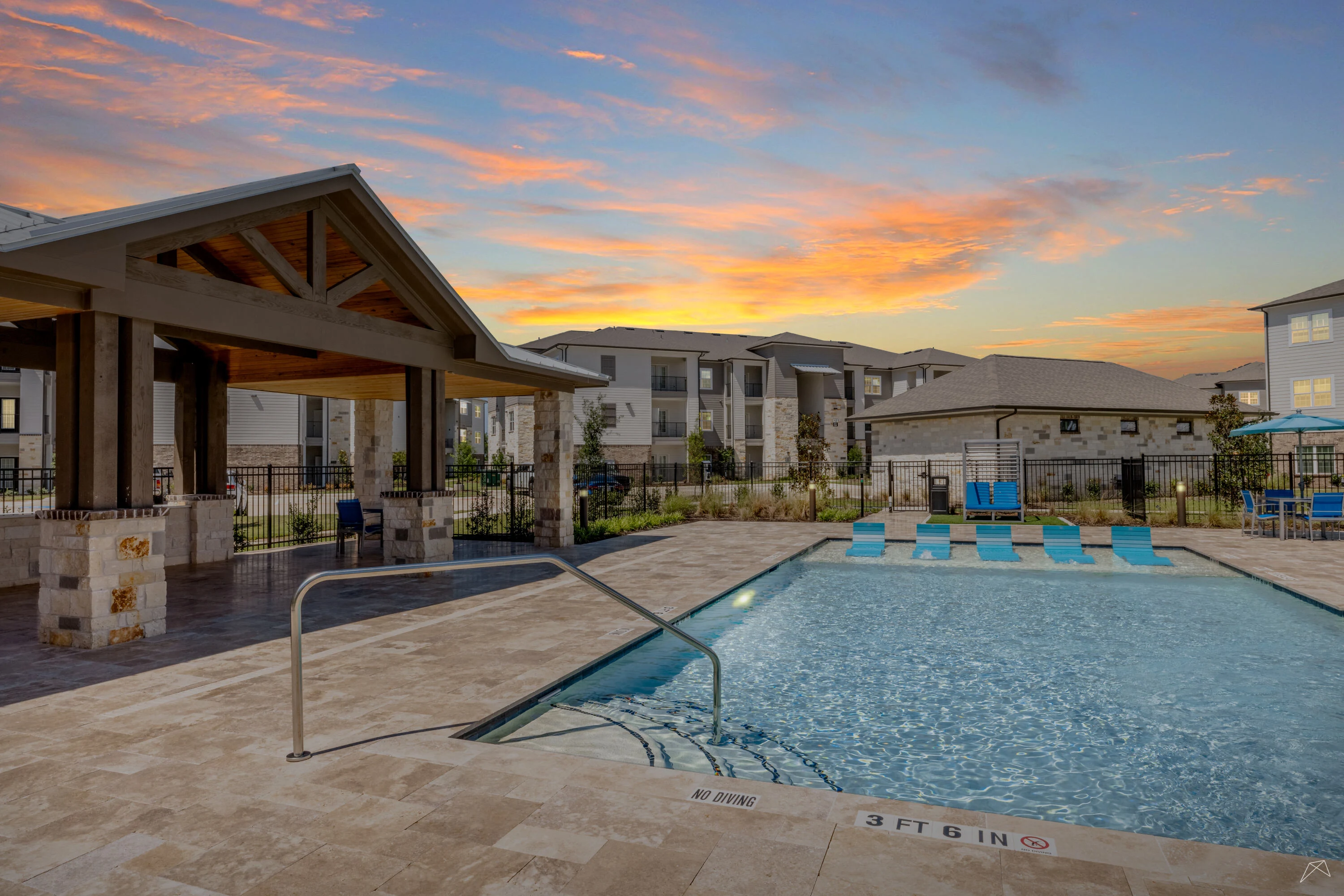 Apartment complex pool area at sunset with loungers, clubhouse, and vibrant sky.
