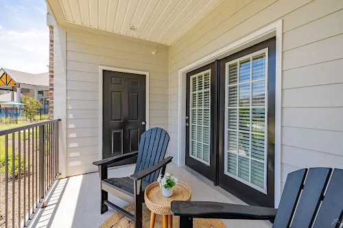Cozy apartment balcony with two dark chairs, a small table, and doors leading indoors, surrounded by greenery.