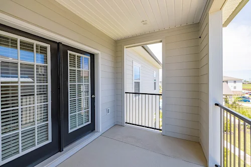 Spacious apartment balcony with double doors and metal railing, featuring a view of neighboring units and clear skies.