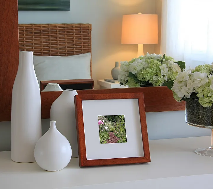 Elegant bedroom corner with decorative white vases, framed art, and fresh hydrangeas reflecting in a wooden mirror frame.