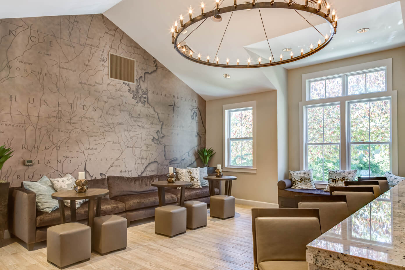 A cozy lounge with a map mural wall, brown sofa, ottomans, small tables, and a large circular chandelier. Sunlight streams through big windows, brightening the space.