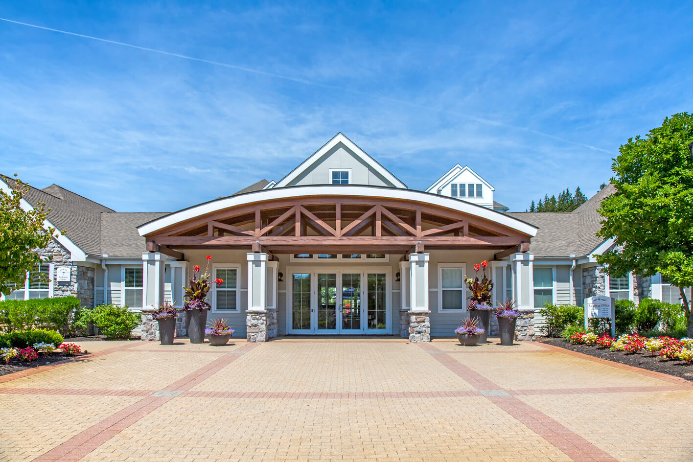 A modern building with a large wooden entrance canopy, stone columns, potted plants, and glass doors, surrounded by landscaped flower beds on a sunny day.