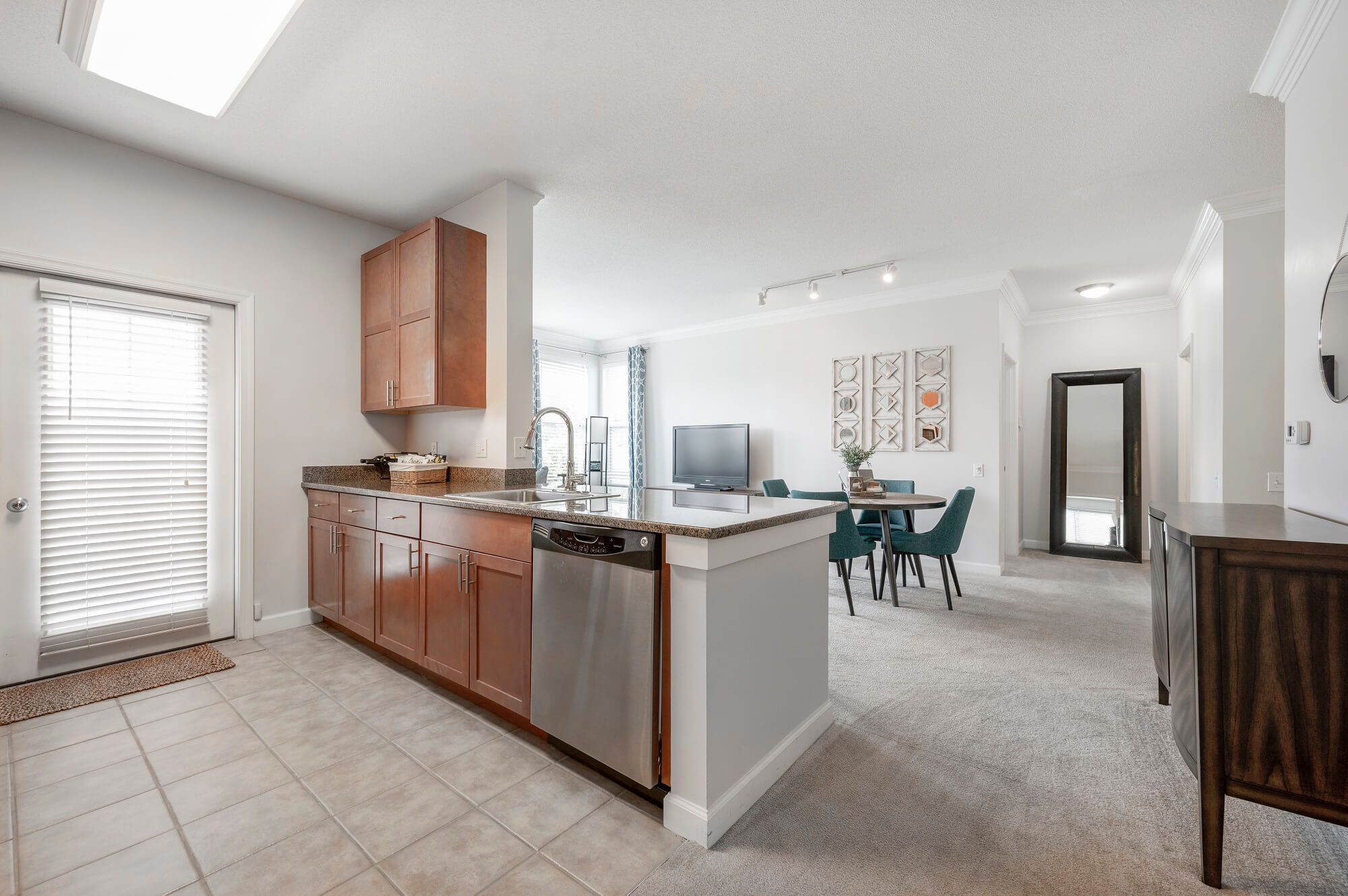 Modern open-concept kitchen with wooden cabinets and stainless steel appliances, adjoining a carpeted dining and living area with teal chairs, a TV, wall art, and large windows letting in natural light.