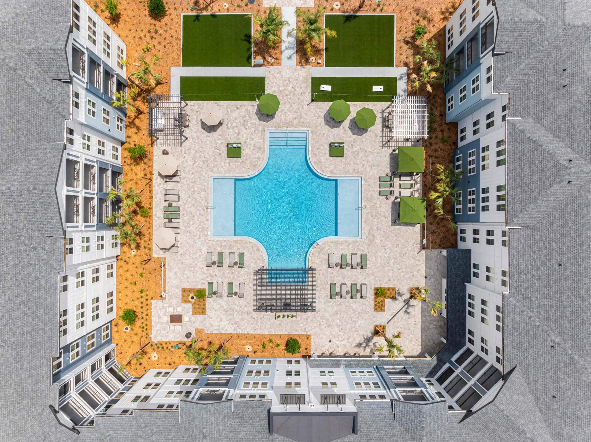 Aerial view of a rectangular swimming pool with lounge chairs, umbrellas, and green spaces, surrounded by a multi-story apartment complex. Explore Apela House: Schedule your tour today and experience resort-inspired living firsthand.