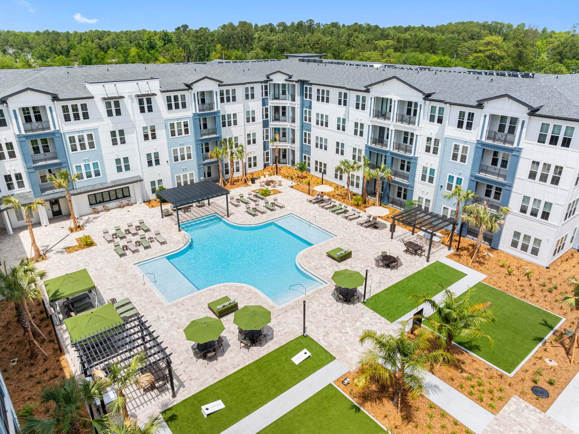 Aerial view of a modern apartment complex courtyard featuring a central swimming pool, lounge chairs, shaded seating areas, and landscaped green spaces with palm trees. Explore Apela House: Schedule your tour today.