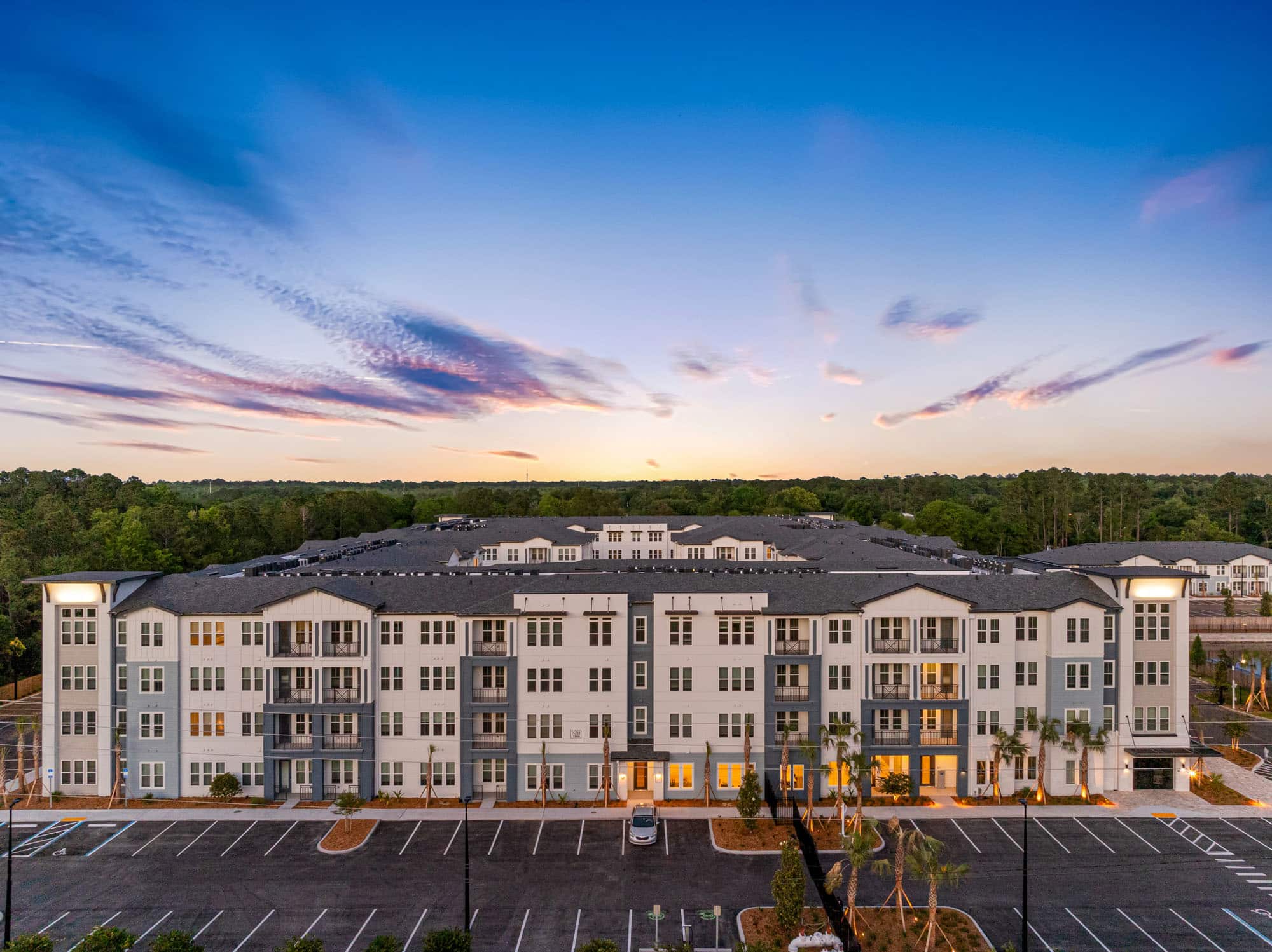 Explore Apela House: Schedule your tour today of this wide, modern apartment building featuring multiple floors, balconies, and an empty parking lot, all set against a sunset sky and surrounded by trees.