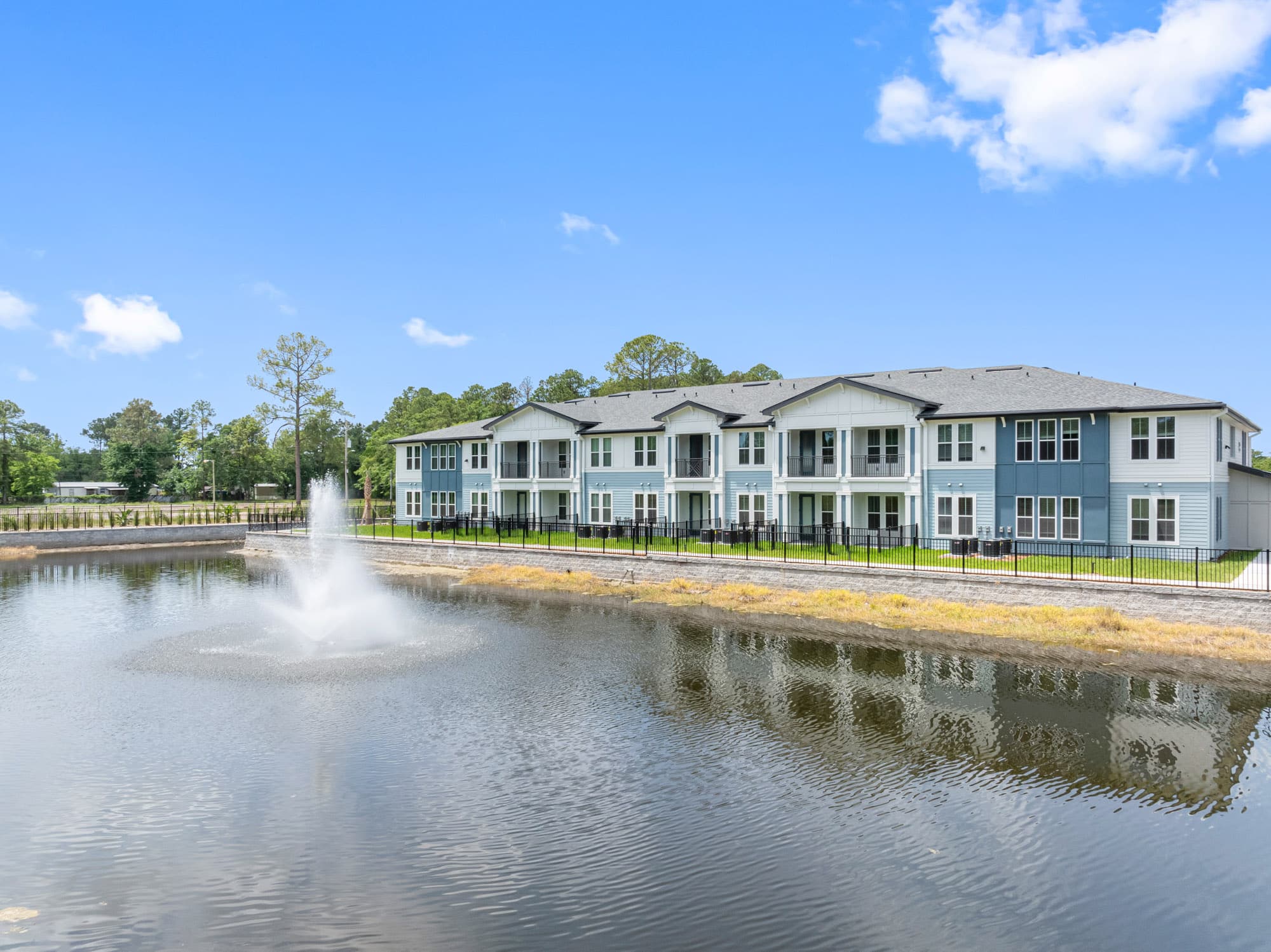 Explore Apela House: A two-story apartment building with balconies stands behind a pond with a water fountain, surrounded by trees and a clear blue sky. Schedule your tour today and discover your new home!.