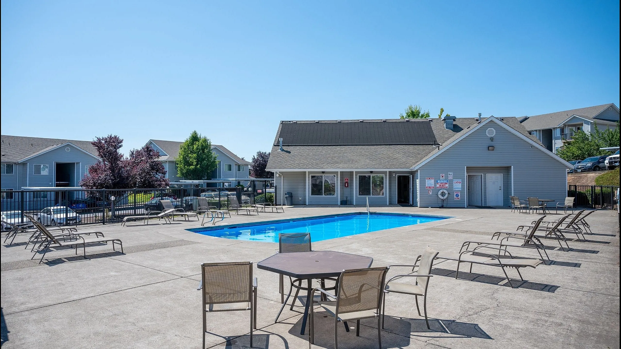 Inviting outdoor pool area with modern lounge chairs and table, surrounded by charming residential buildings under a clear blue sky.