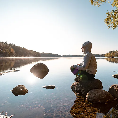 Serene lakeside view with a person meditating on rocks, surrounded by autumn trees and calm reflections.
