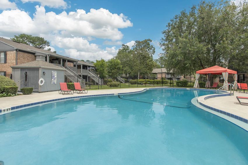 A clear blue swimming pool surrounded by lounge chairs and green landscaping. In the background, there are apartment buildings and a canopy structure. The sky is partly cloudy, creating a bright and inviting atmosphere.