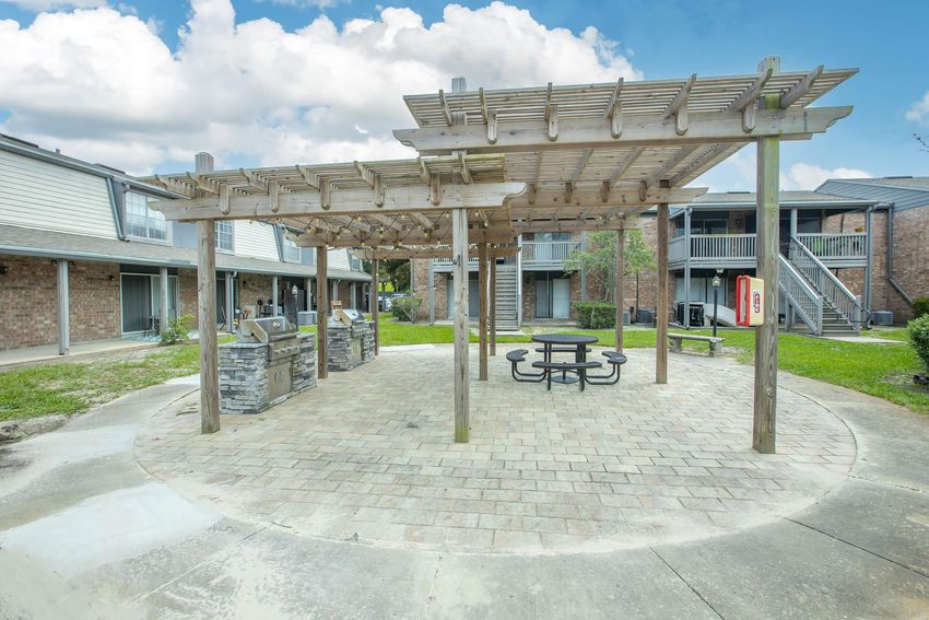 Outdoor communal area with a pergola, seating at a circular stone patio, and two barbecue grills. Surrounding structures are residential buildings with brick exteriors and balconies. The sky is partly cloudy, creating a bright atmosphere.