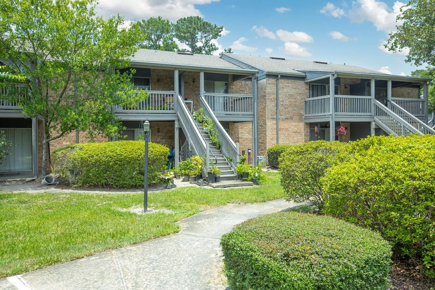 A well-maintained two-story apartment building with brick exterior, featuring several balconies and stairs leading to the entrances. Lush green landscaping surrounds the walkways and steps, with bushes and trees providing a pleasant outdoor environment on a sunny day.
