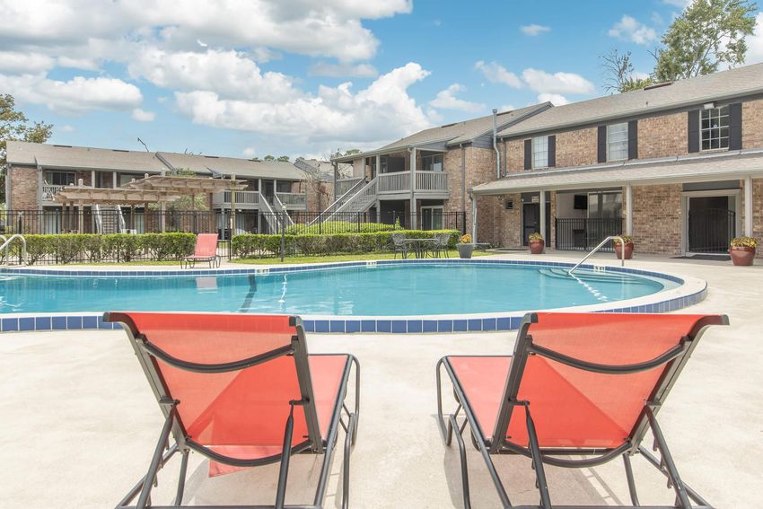 A serene outdoor pool area with two red lounge chairs in the foreground. The pool is surrounded by greenery and a wooden pergola. In the background, there are two multi-story brick buildings with balconies, under a partly cloudy blue sky.