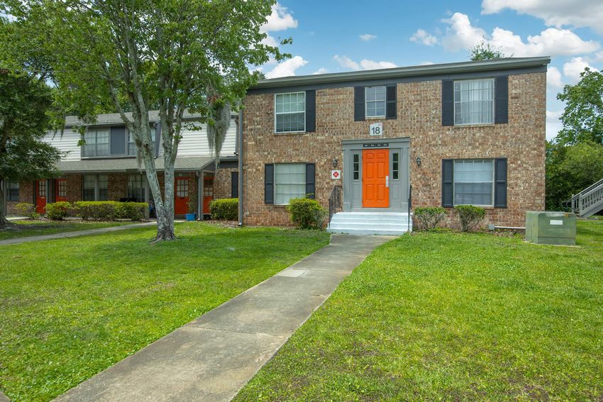 A two-story brick apartment building with multiple units. The front features an orange door and white steps, flanked by green grass and shrubbery. Pathway leads to the entrance. Nearby, there are additional units with red doors, surrounded by trees and a clear blue sky.