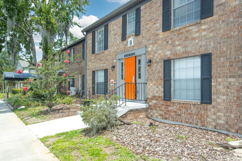 A brick apartment building with an orange door, featuring green landscaping, trees, and flowering plants. The entrance has a set of steps leading up to the door, and there are window shutters on either side of the windows. The scene is bright with a clear blue sky.