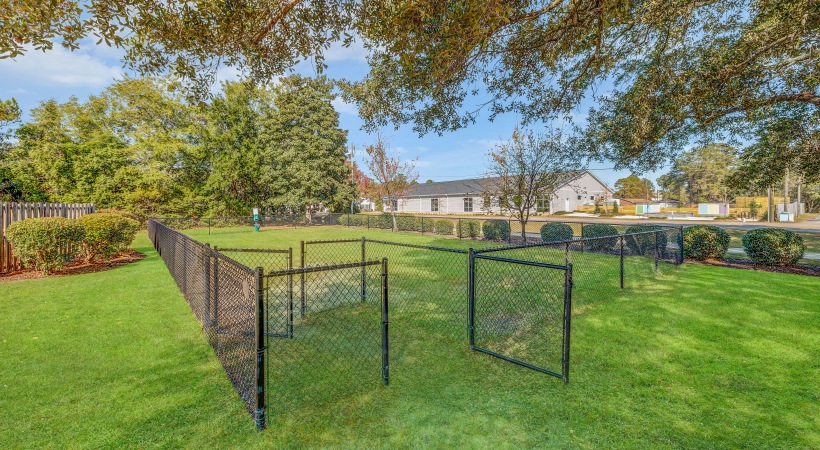 Leland, NC, Pet-Friendly Apartments - Flats at Leland - Outdoor, Gated Off-Leash Dog Run in a Large Grassy Area with a Pet Waste Sanitary Station, Surrounded by Trees and Other Greenery