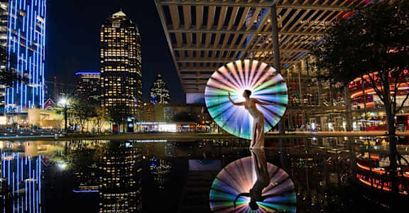 A large CD-shaped sculpture is reflected in the water in front of a city skyline at night.
