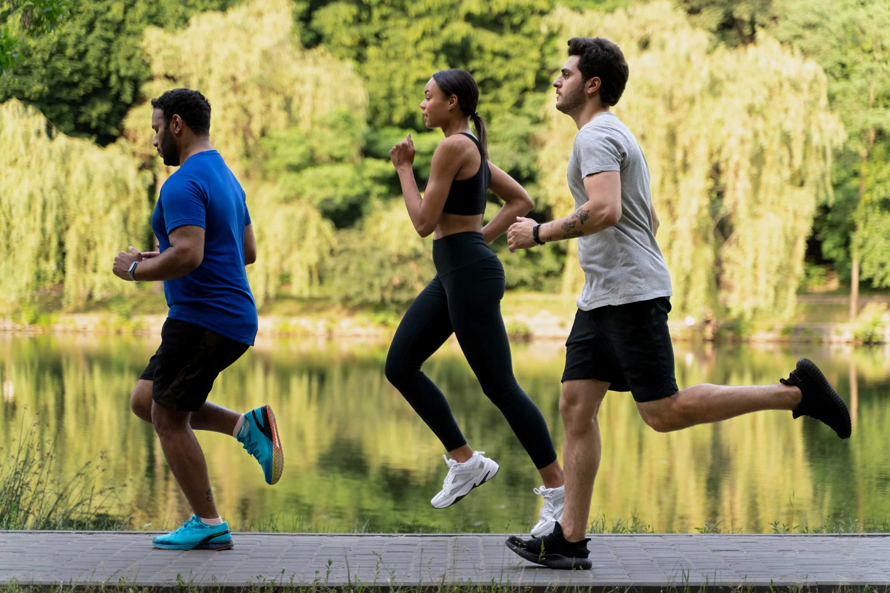 Three individuals running along a pathway by a lake surrounded by greenery. Two men and one woman are jogging, wearing athletic clothing. The background consists of trees and a reflective water surface.