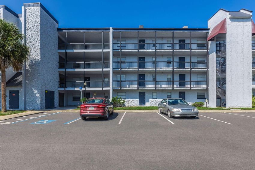 A view of a three-story residential building with a textured gray exterior. The building features multiple balconies and staircases, with a parking lot in the foreground. Two cars, a red sedan and a silver sedan, are parked in designated spaces. The sky is clear and blue, indicating a sunny day.