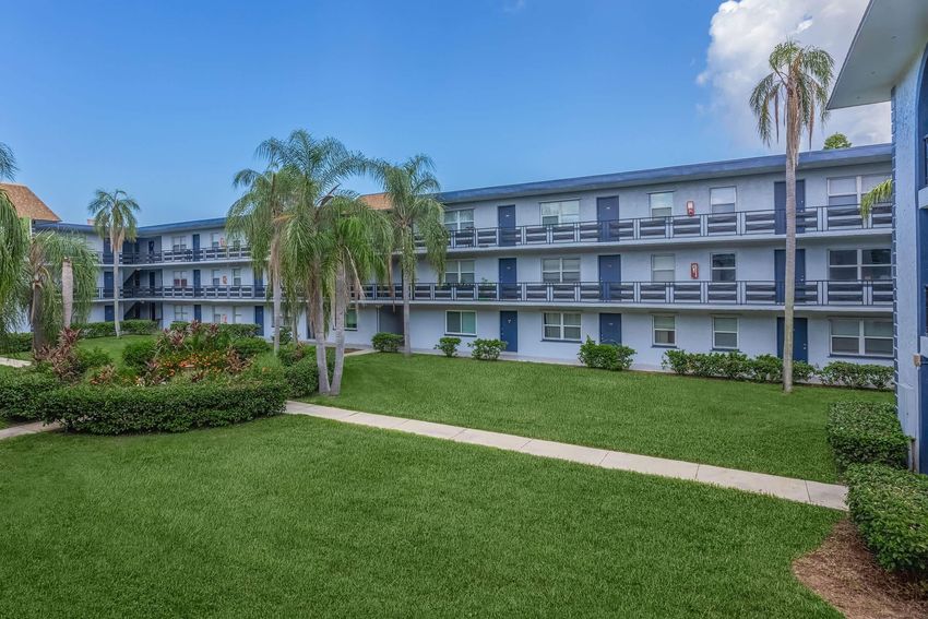 A view of a multi-story apartment building surrounded by palm trees and well-maintained grass. The structure features multiple rows of balconies, with a clear blue sky in the background. Pathways connect the entrances to the building, creating a serene, inviting environment.