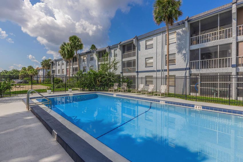 A clear blue swimming pool surrounded by a gray concrete deck, with lounge chairs and palm trees in the background. Several multi-story residential buildings with balconies are visible behind the pool, under a bright sky with fluffy clouds.