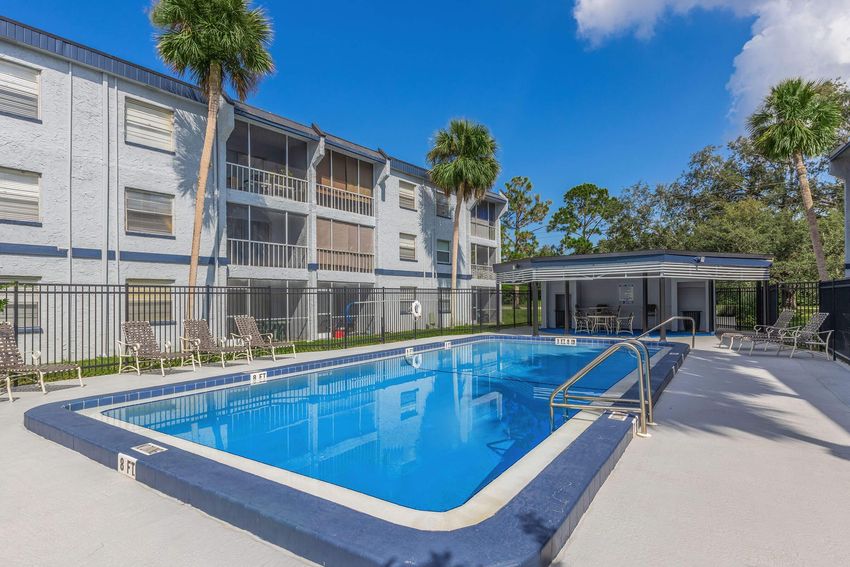 A clear blue swimming pool surrounded by a gated area, with lounge chairs positioned nearby. In the background, there are multi-story apartment buildings painted in light blue, with palm trees and green foliage framing the scene under a bright blue sky with a few clouds.