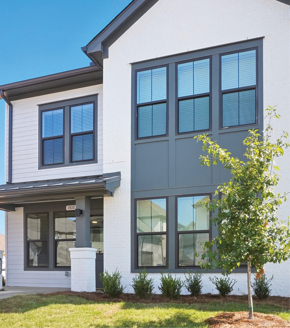 Modern two-story house with white brick exterior, black framed windows, and small front yard tree against clear blue sky.