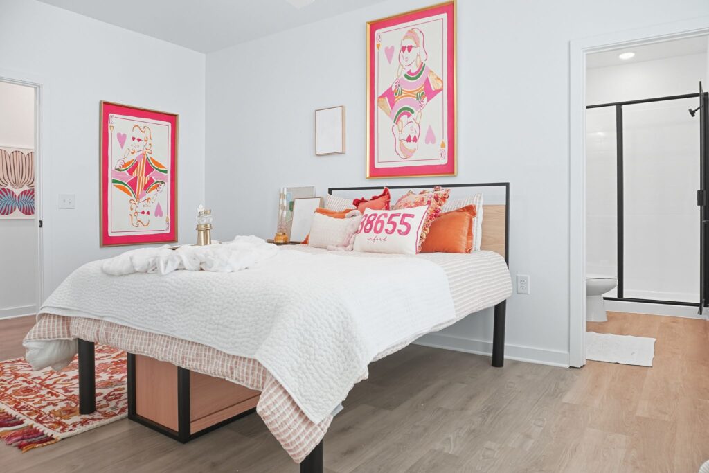 Modern bedroom with colorful art, white bedding, and decorative pillows, next to a stylish bathroom.