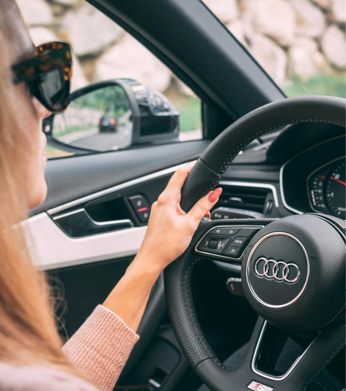 Person driving an Audi, holding steering wheel with scenic road visible in side mirror.