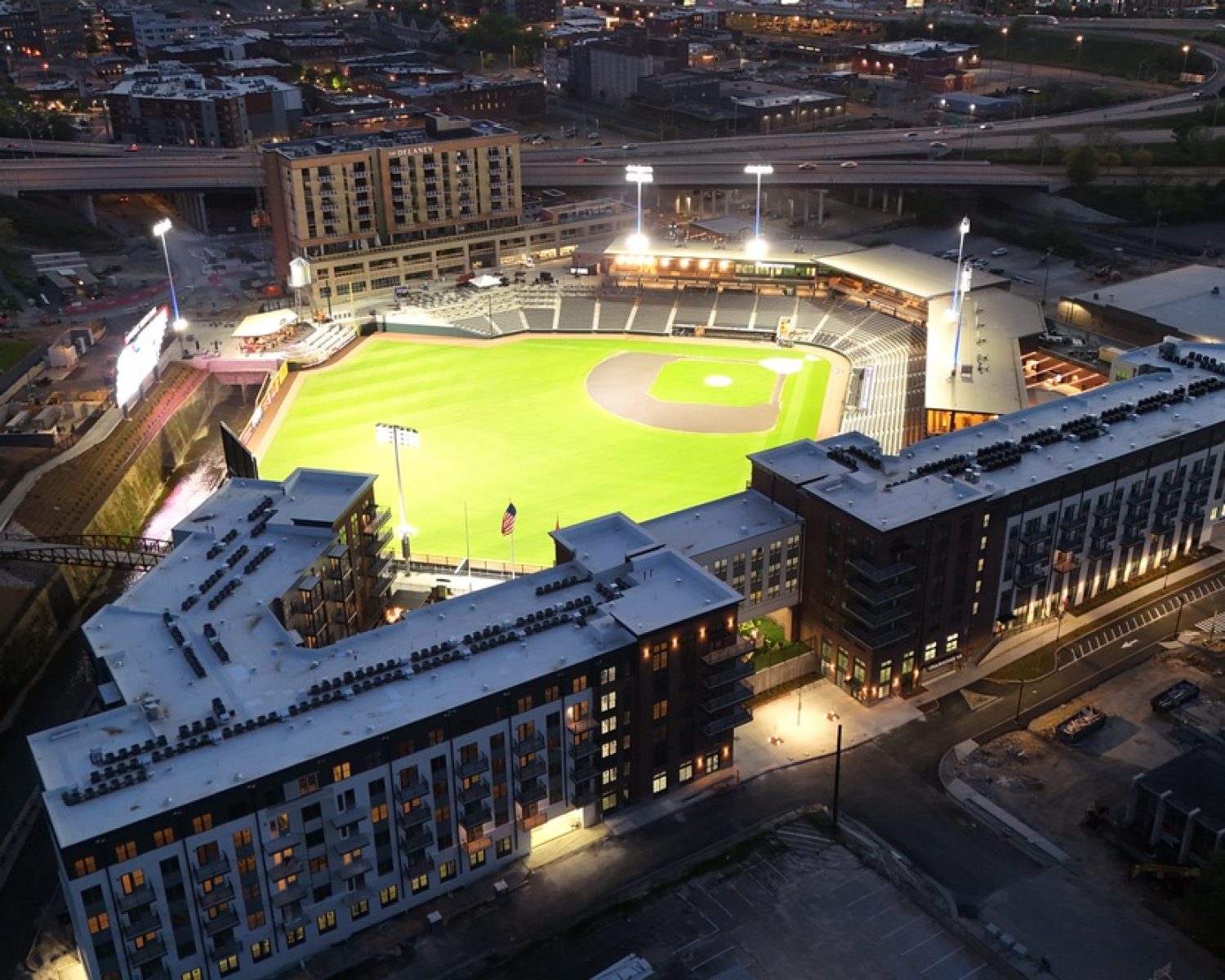 Apartments For Rent Downtown Knoxville Aerial view of a brightly lit baseball stadium surrounded by buildings and roads at dusk, with the cityscape visible in the background. Knoxville Apartments