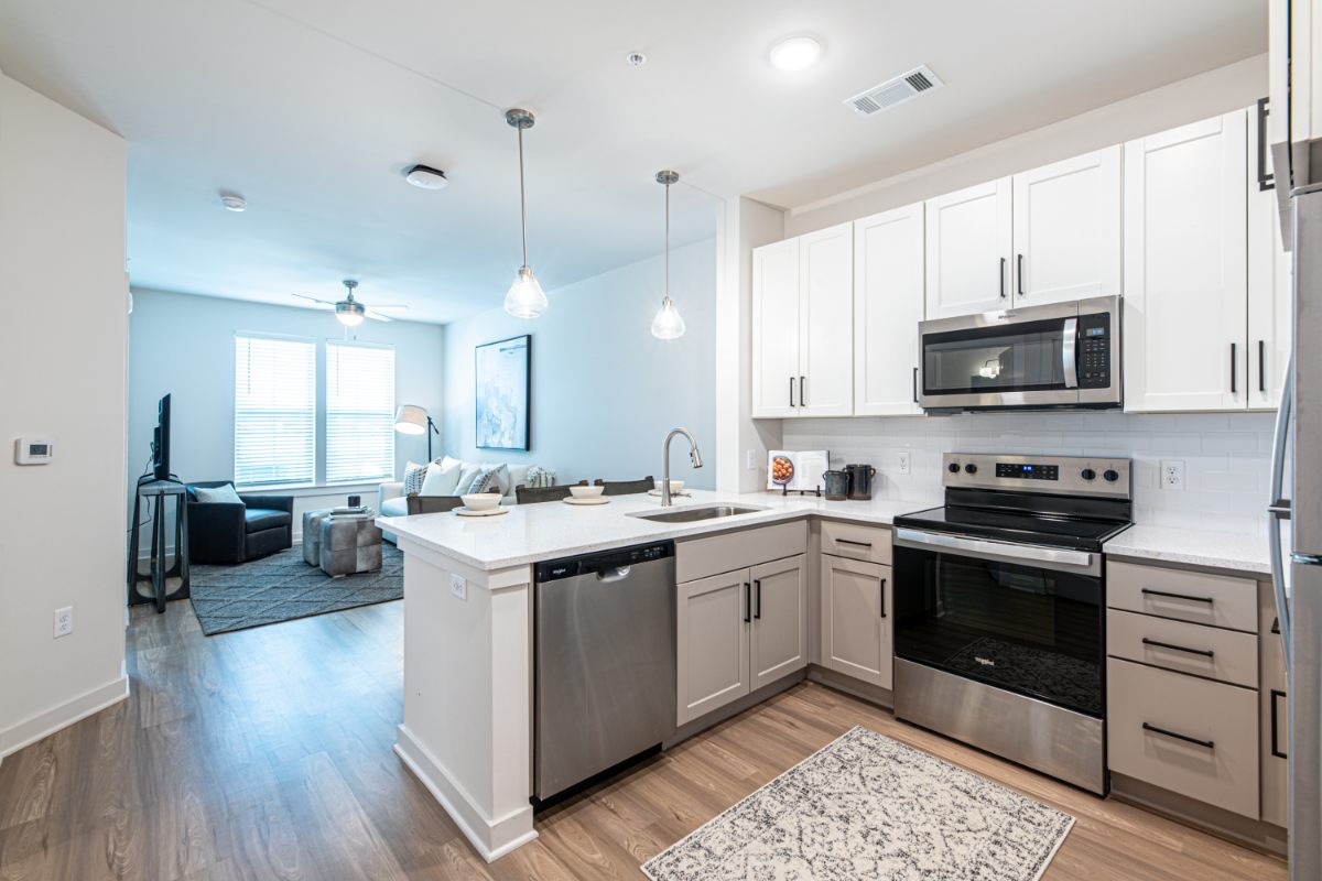 Studio Apartments Apartments For Rent Downtown Knoxville Modern kitchen with white cabinets, stainless steel appliances, and an island, opening to a living room with large windows and a gray sofa. Knoxville Apartments