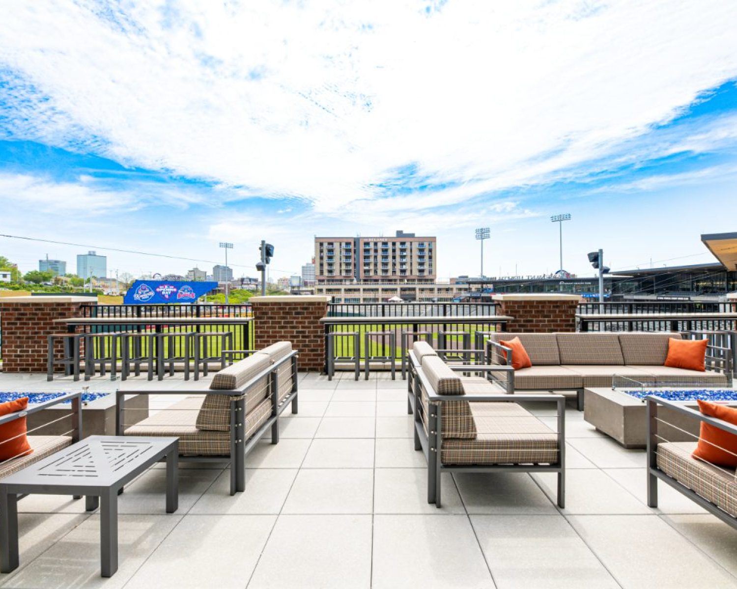 Apartments For Rent Downtown Knoxville Rooftop patio with modern outdoor seating, fire pits, and a view of a baseball stadium and city buildings under a blue sky. Knoxville Apartments