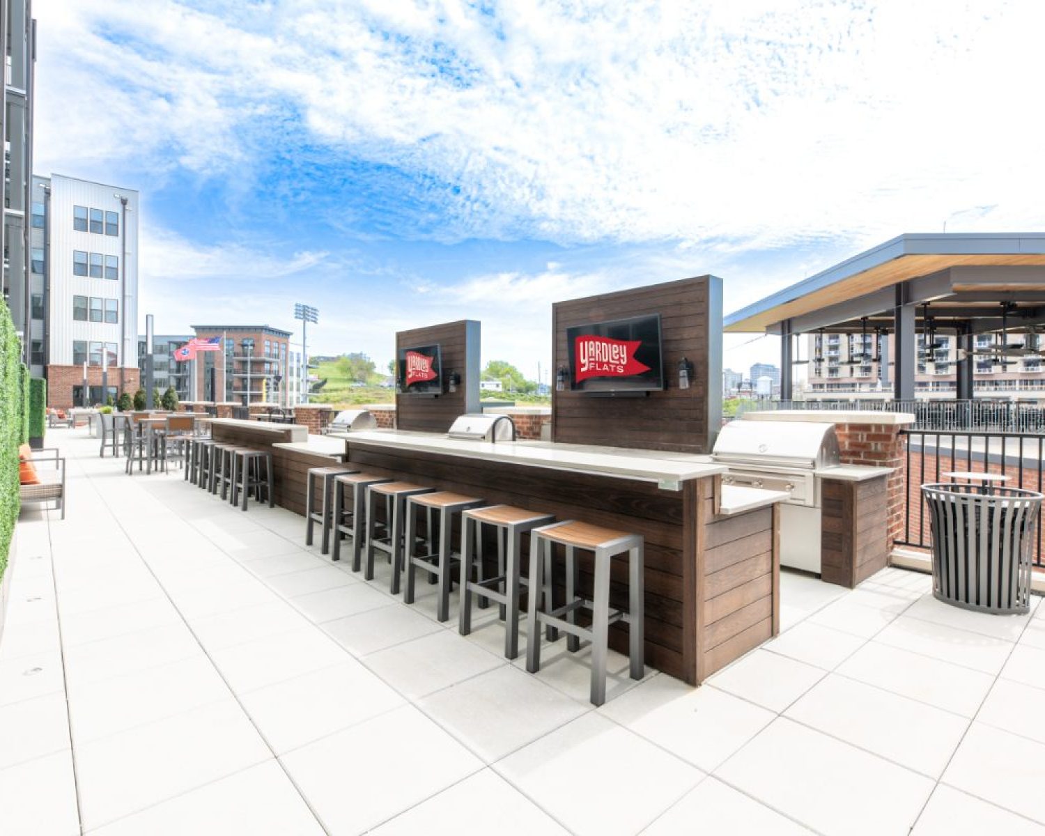 Apartments For Rent Downtown Knoxville Outdoor rooftop patio with bar seating, built-in grills, mounted TVs, and cityscape views under a partly cloudy sky. Knoxville Apartments