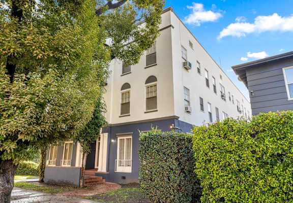 A white building with a grey door and windows is surrounded by greenery.