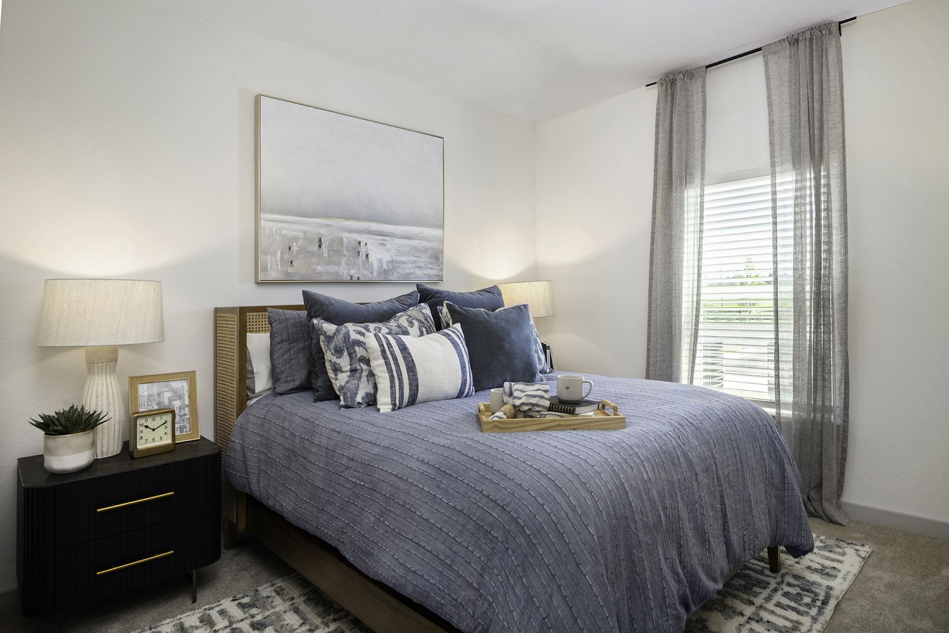 Cozy bedroom with a gray bed, black nightstand, lamp, wall art, and sheer curtains at Livano Wildwood.
