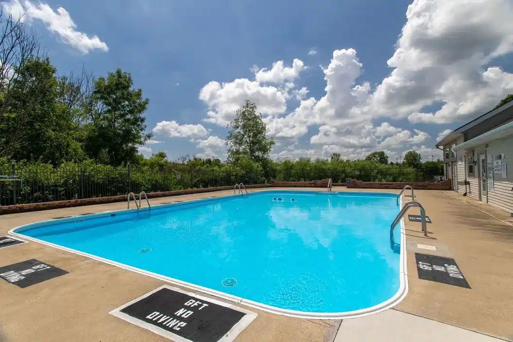Outdoor swimming pool with clear blue water, surrounded by a concrete deck, metal railings, and a "5 FT NO DIVING" sign, with trees and clouds in the background.