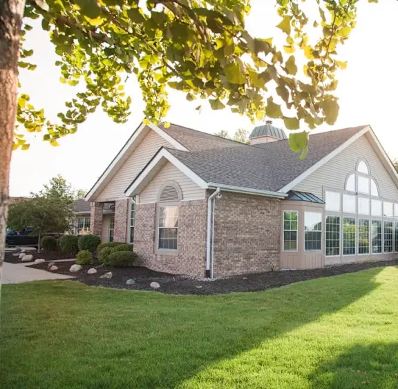 A single-story brick building with large windows is surrounded by green grass, shrubs, and trees on a sunny day.