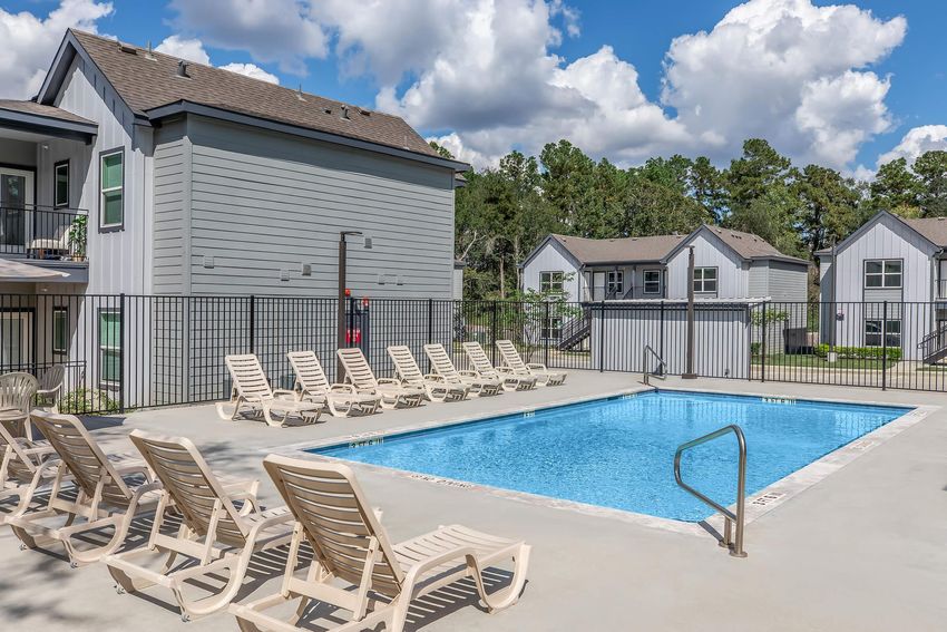 A clear swimming pool surrounded by lounge chairs, with a fence around the pool area. In the background, there are two multi-story apartment buildings and a sunny sky with scattered clouds. The setting is serene and ideal for relaxation.