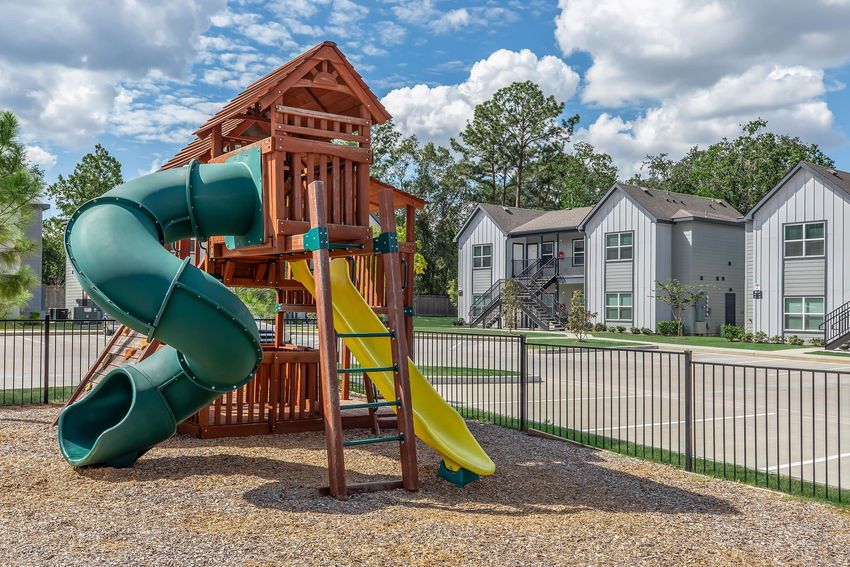 A colorful playground with a wooden structure featuring a green slide and a yellow slide, set in a gravel area. In the background, there are two modern residential buildings surrounded by greenery and trees, under a partly cloudy sky.